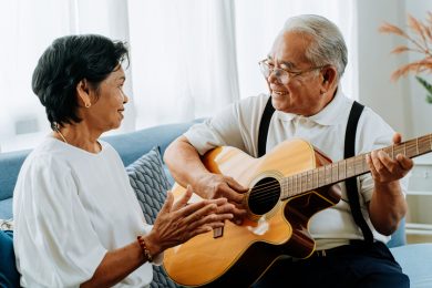 Asian senior couple sitting on the sofa and playing acoustic guitar together. Happy smiling elderly woman clapping hands while old 70s guitarist husband singing. Enjoying retirement life at home.